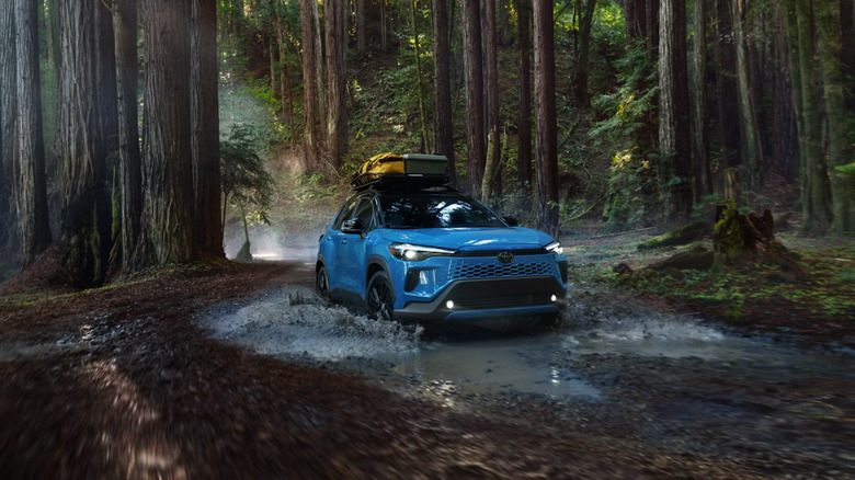 A dark blue 2026 Toyota Corolla Cross SUV splashing through a puddle in the middle of a shaded forest.
