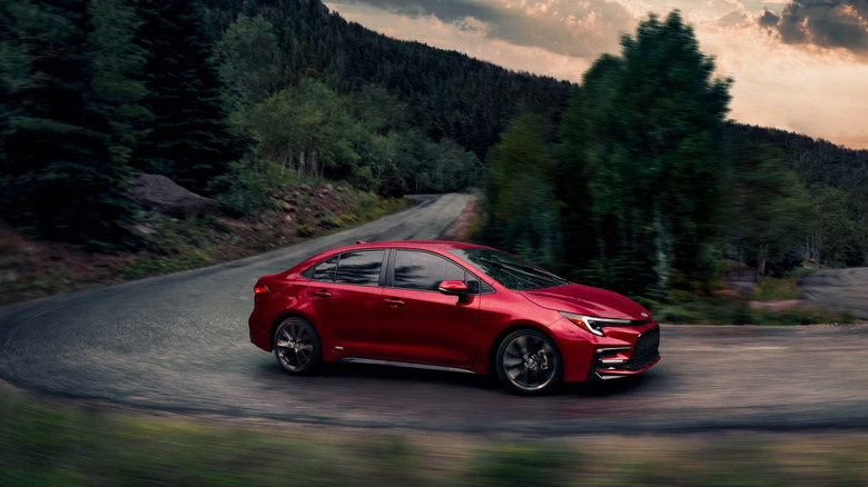 A red metallic 2026 Toyota Corolla taking a turn at speed down a forested, woody road in the evening.