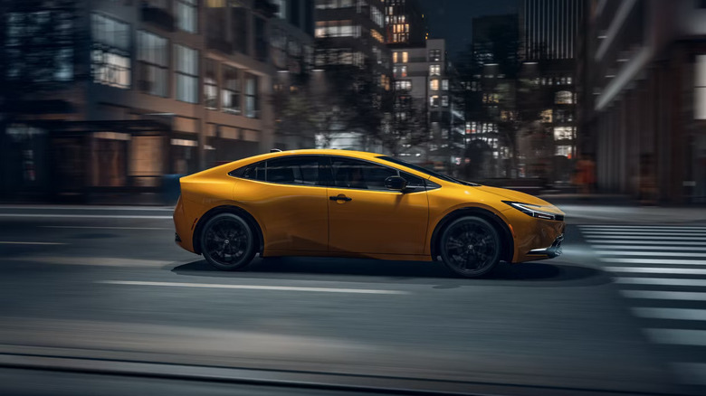 A yellow 2026 Toyota Prius HEV photographed here as it moves through a city street at night, with the backdrop blurred.