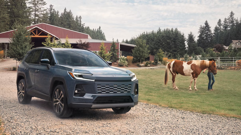 A blue-gray 2026 redesigned RAV4 standing on a gravel path, with a woman leading a horse on grass in the back.