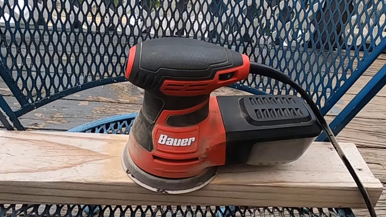 The Bauer 2.8 amp palm sander sitting on a half-sanded piece of wood laid across a chair.