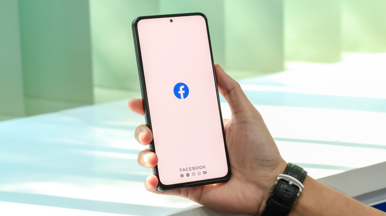A person holds a phone which is displaying te Facebook logo on a white background.