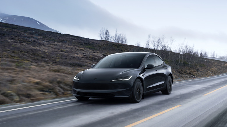 A dark Tesla Model 3 on a wet rural road with misty hills in the distance