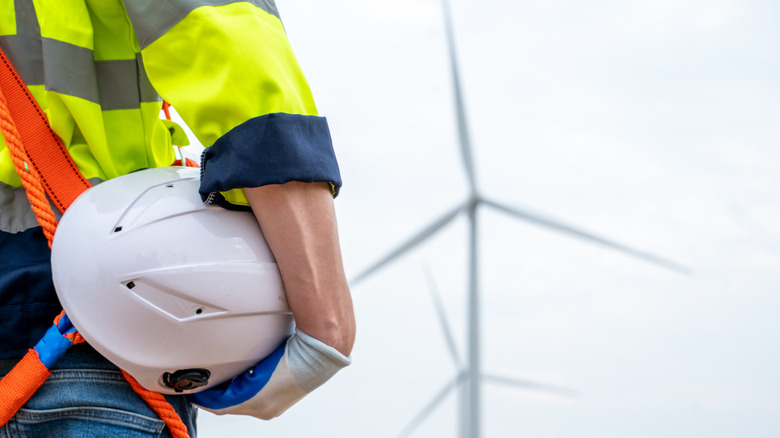 Close-up of a man in a yellow safety shirt and orange harness holding a white hard hat near several large wind turbines
