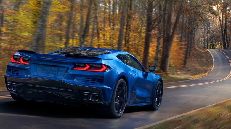 Rear view of blue 2026 Chevrolet Corvette E-Ray driving down a forested two-lane road