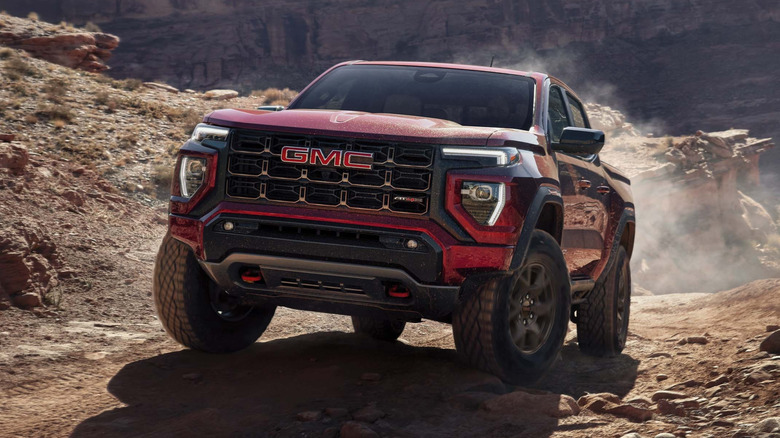 Front 3/4 view of 2027 GMC Sierra 1500 pickup driving off road in mountainous, rocky setting