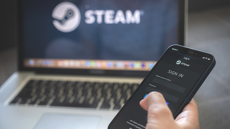 Person holding an iPhone with Steam app open with a MacBook in the background