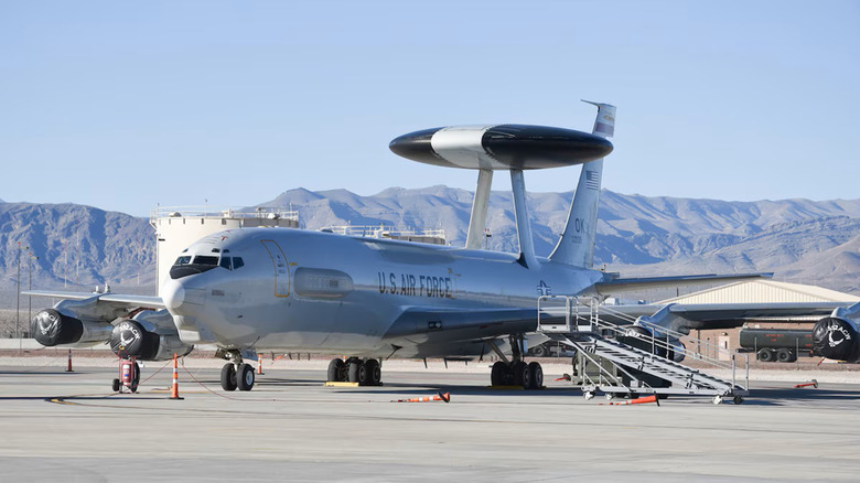 Silver plane with large radar dish on top and U.S. Air Force written on the side, parked on an airfield with steps. Moutains are in the background.