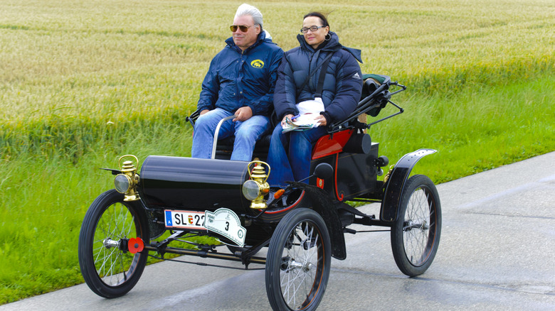 Man and woman wearing winter coats riding in 1902 Oldsmobile Curved Dash runabout on country road
