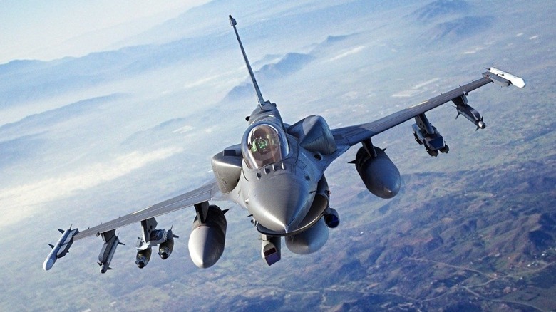 F-16 fighter jet flying over partially cloudy skies with visible terrain below.