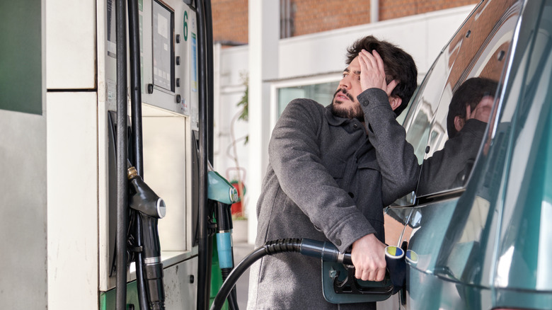 A man in a gray coat looking worried while he fills up his vehicle at a gas pump