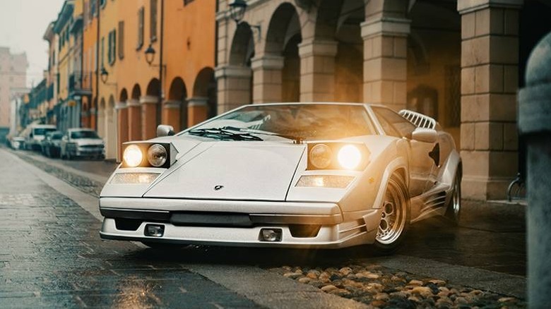 A silvery-white Lamborghini Countach from the 1970s parked in front of an old building with its headlamps on.