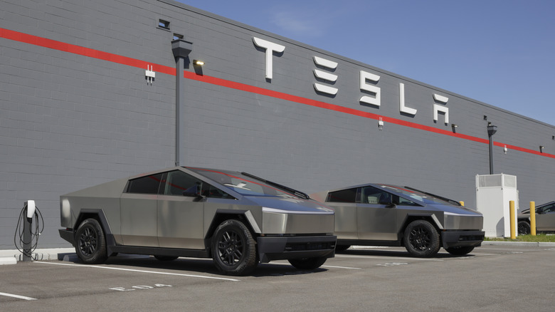 Two silver Tesla Cybertrucks parked outside a large gray building with the Tesla logo on it.