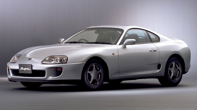 A silver 1993 Toyota Supra standing against a gray studio back drop, front-left three-quarter view visible.