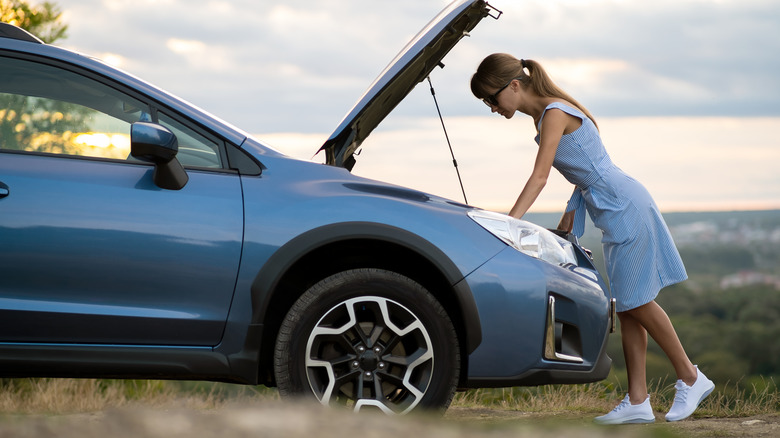 Person looking under the hood of a broken-down blue Subaru crossover parked on side of the road