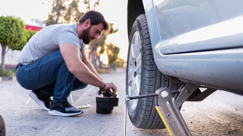 A bearded man in a gray shirt and jeans replaces a flat tire on the side of the road