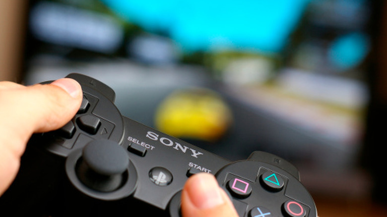 Close-up of hands holding a Sony DualShock 3 controller with an out-of-focus videogame in the background.