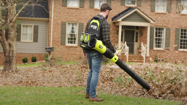 Man with Ryobi battery backpack leaf blower blowing leaves in front of large brick house