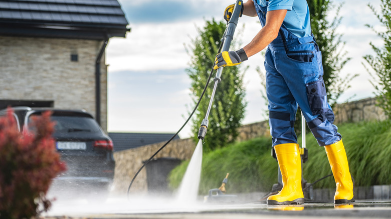 A homeowner wearing a safety floves and boots using a powerful pressure washer to clean a dirty driveway