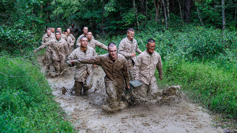 A group of soldiers walking in mud with the two at the front holding an ammo case together.