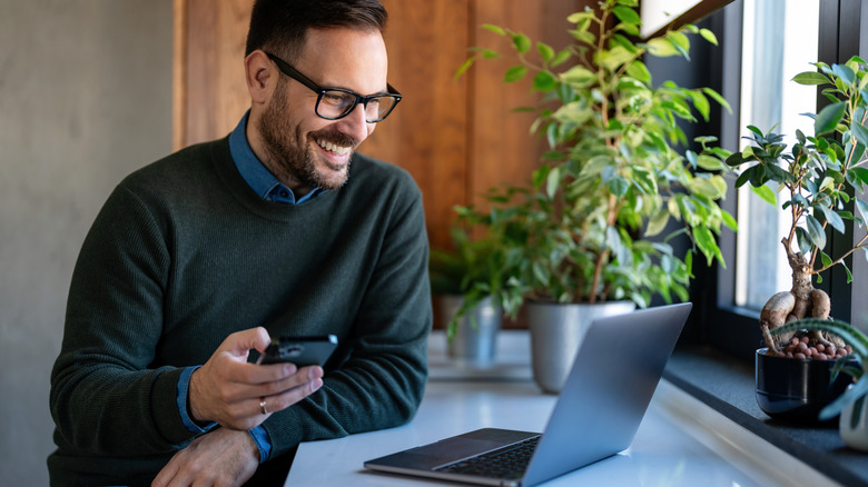 Happy man using smartphone and smiling at laptop screen