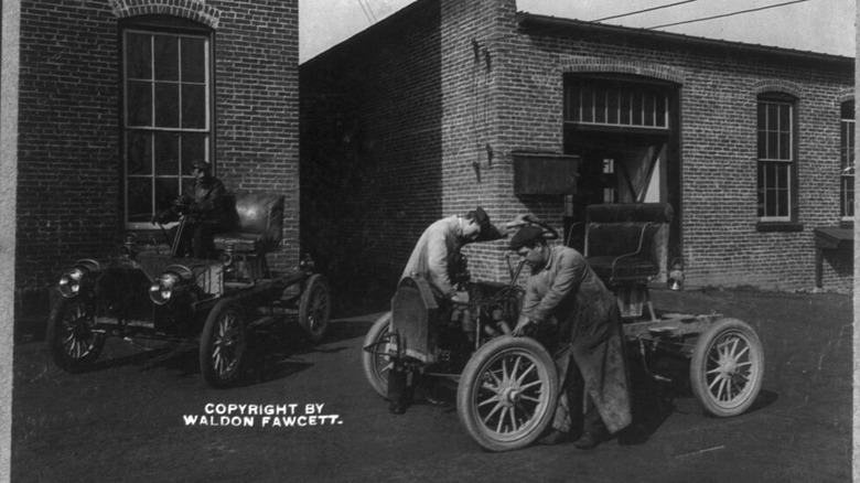 Two men working on an automobile outside of a garage circa 1906.