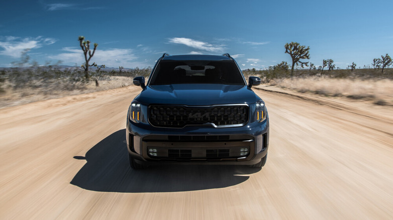 2025 Kia Telluride driving toward the camera along a dirt road with blue sky and trees in the background
