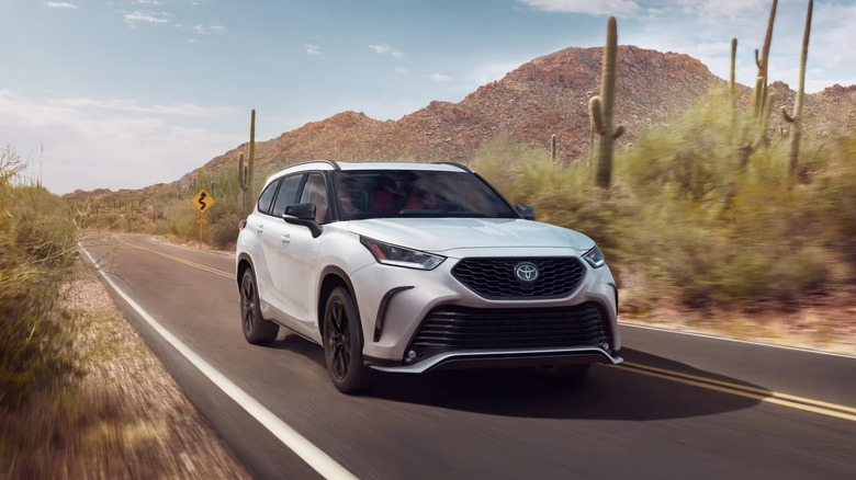 White 2025 Toyota Highlander driving on a highway toward the camera with cactus and shrubs in the background