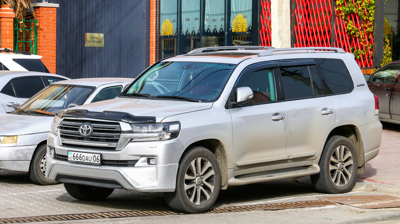 A white Toyota Land Cruiser 200 parked on a city street surrounded by other cars in the afternoon.