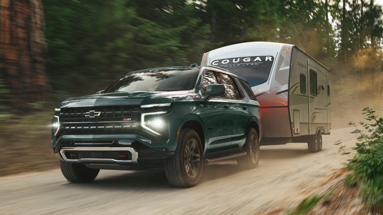 Front 3/4 view of green 2026 Chevy Tahoe towing trailer down dirt road.