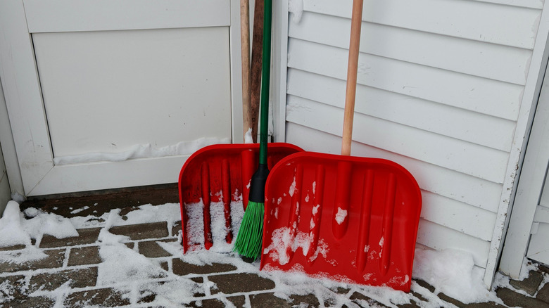 Two bright red snow shovels and a green broom sitting on a porch leaning against a white house