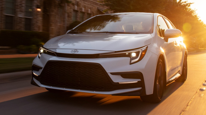 Front 3/4 view of Toyota Corolla driving in urban setting, backlit by sunset