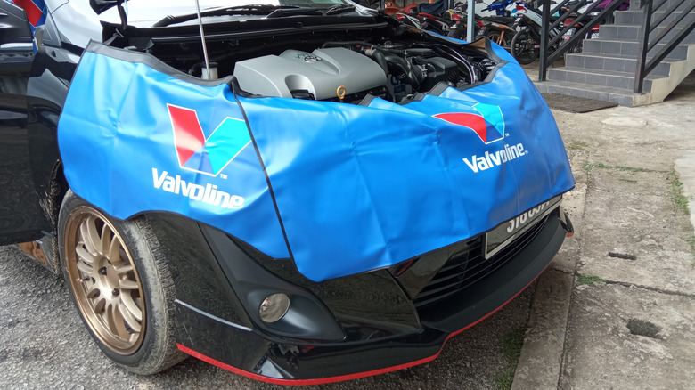 A Toyota hatchback with the hood open and protective Valvoline-branded covers around the front fender and bumper.