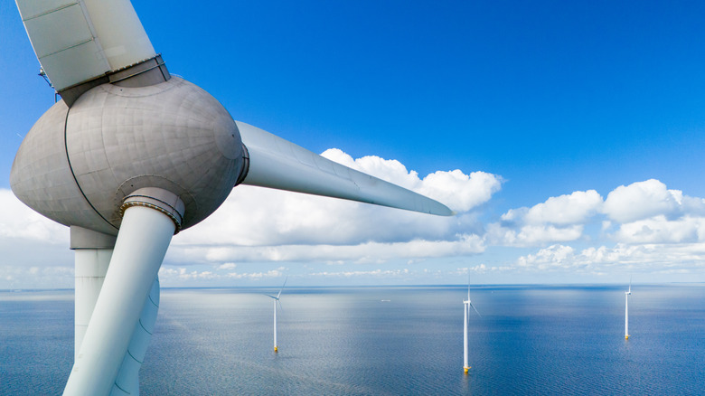A close-up of an offshore wind turbine with additional turbines and blue sky and clouds in the background