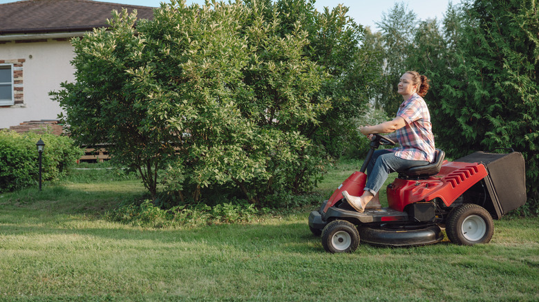 A middle age farmer women mows the lawn with a ride-on lawn mower near a house.
