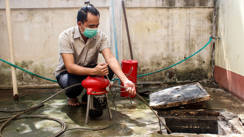 Man with mask clearing an outdoor drain pipe with an electric drain auger