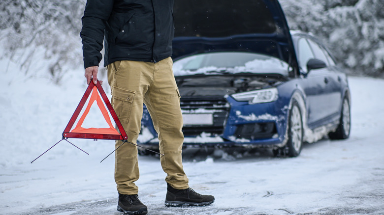 Person in snowy terrain carrying red emergency warning triangle with broken-down Audi in background