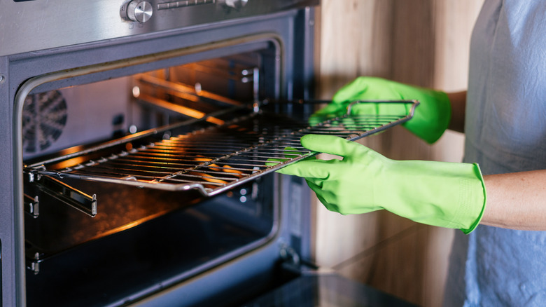 a person wearing green rubber gloves taking out a dirty oven rack from an oven