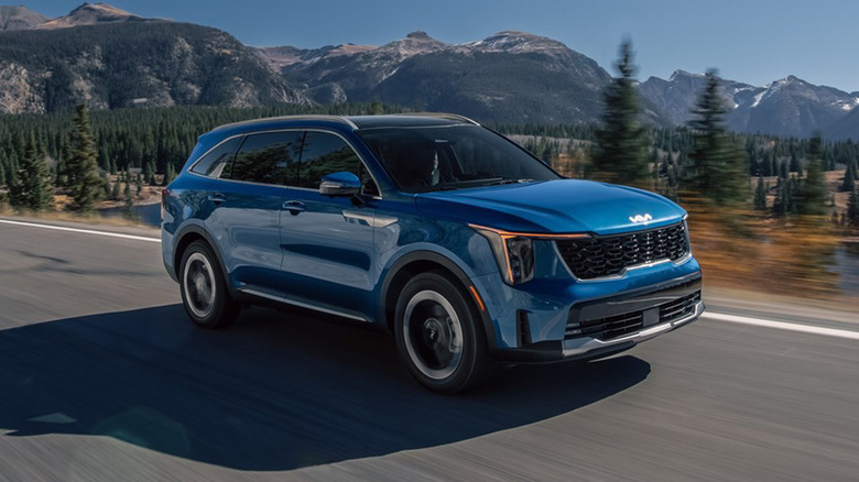 Blue 2025 Kia Sorento SUV driving through a scenic mountain highway with pine forests and lake views