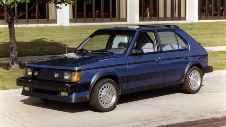 Front 3/4 view of blue Dodge Omni GLH parked in front of building and grass.
