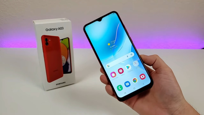 A person holding a Samsung A03 phone in front of the original box on a white table