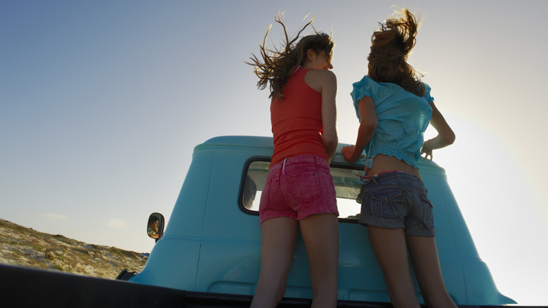 Two girls stand on the back of a blue pickup truck while moving