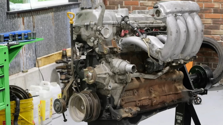 A Toyota 3RZ-FE engine on an engine stand inside a car workshop, with a brick wall and tools visible in the back.