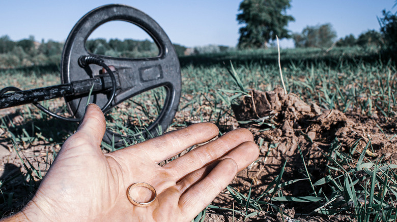 A man's hand holding a gold ring with a metal detector in the grass in the background