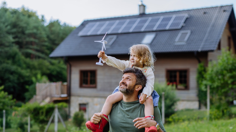 Father carrying his daughter on his shoulders near their family home with solar panels on the roof.