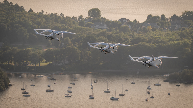 Three electric BETA Technologies airplanes in the air over a body of water filled with sailboats