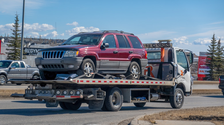 A towing flatbed truck transports a maroon Jeep Grand Cherokee Laredo on the street