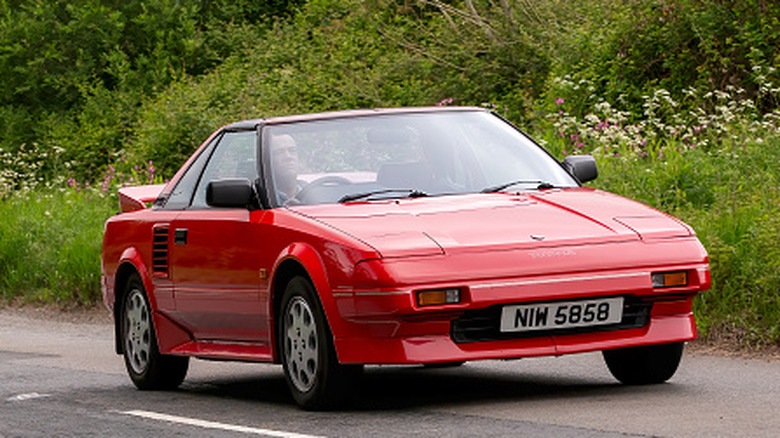 A right-hand drive red 1991 Toyota MR2 on a country road, greenery in the background