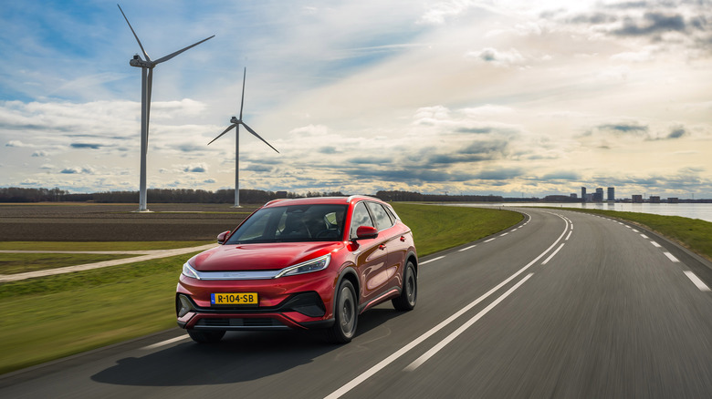 BYD ATTO3 Road in red driving on a clear road with a cloudy sky and wind turbines in the background.