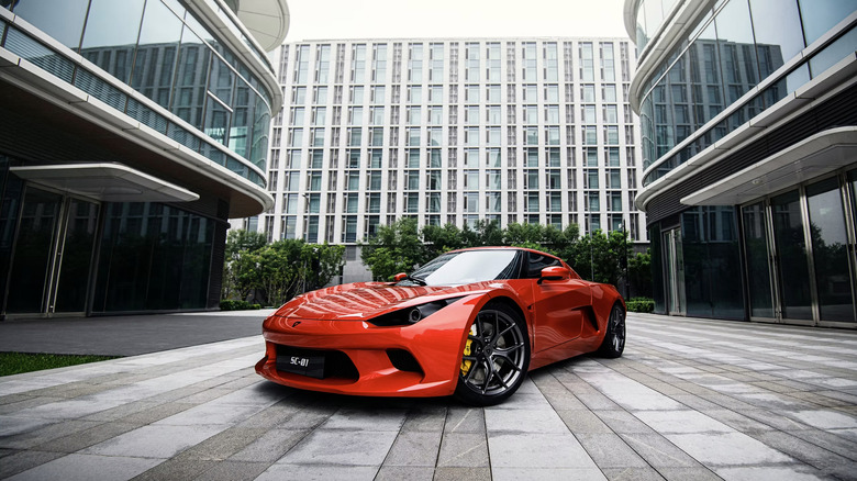 A red SC01 car parked on paving between two modern, glass-clad buildings